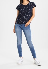 Kvinna iklädd en marinblå t-shirt med fågelmönster, ljusblå skinny jeans och vita plattformssneakers, som står med ena handen i fickan.