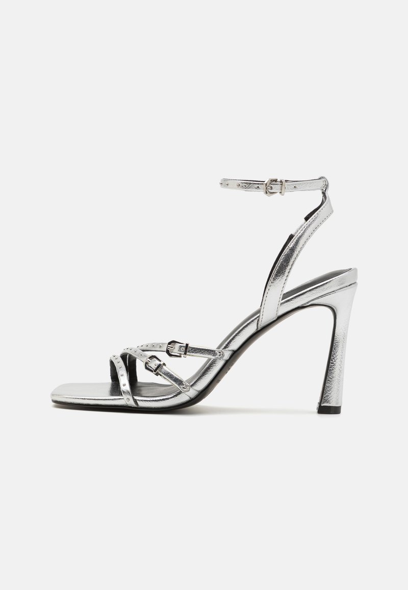 Srebrna metalna sandala na visoki peto z zaponko, ki ima križane trakove z detajli zaponk in gladko, sijočo teksturo.