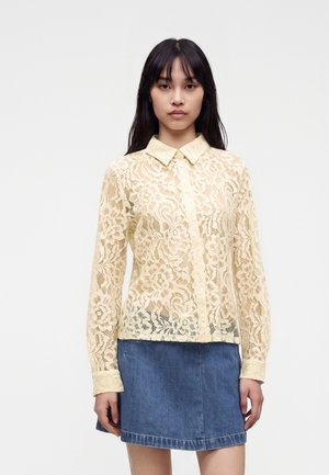 Femme portant une chemise en dentelle florale couleur crème à manches longues avec un col pointu et une jupe en jean bleue, debout devant un fond uni.