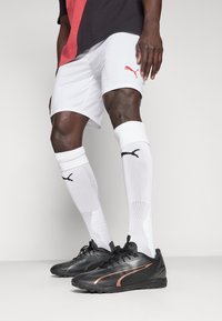 Svarta atletiska skor med strukturerad yta och koppardetaljer, vita shorts med röd logotyp och vita strumpor med svart logotyp.