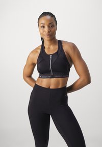 Schwarzer Sport-BH mit Reißverschluss, ausgestattet mit einem kontrastierenden Logo-Bund. Kombiniert mit hochtaillierten schwarzen Leggings, beide aus dehnbarem Material.