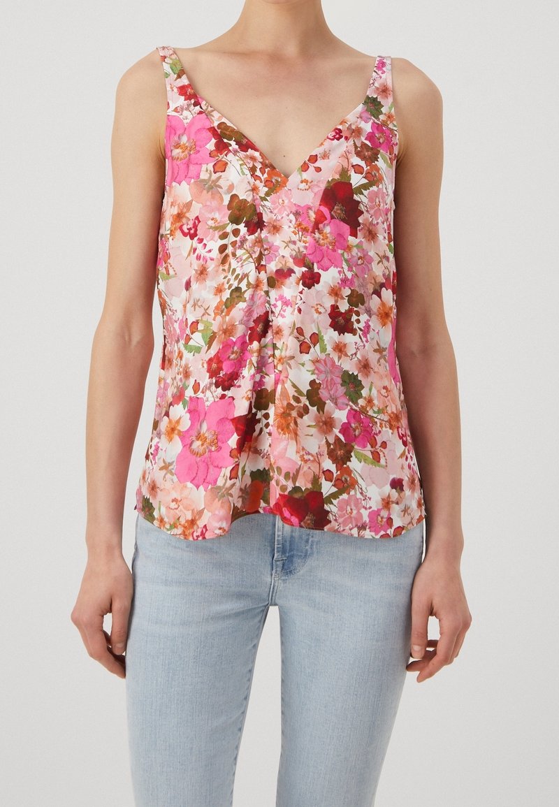 Femme portant un haut sans manches à col en V à motif floral dans des tons roses, rouges et oranges, associé à un jean bleu clair sur un fond uni.