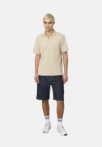 Polo shirt en tricot beige à manches courtes et décolleté en V, associé à des shorts en denim foncé arrivant aux genoux et des baskets blanches avec des rayures bleues sur les chaussettes.