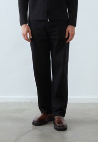 Pantalon noir à coupe décontractée, en tissu robuste avec une coupe droite. Associé à des chaussures en cuir marron et un haut noir zippé.