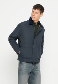 Giacca blu navy con collo e chiusura a scatto, realizzata in tessuto morbido. Presenta una tasca frontale e un dettaglio di fodera testurizzata.