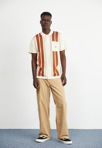 Camisa de manga corta color crema con rayas verticales marrones, un cuello y un bolsillo en el pecho izquierdo, combinada con pantalones de pana beige y zapatillas negras.