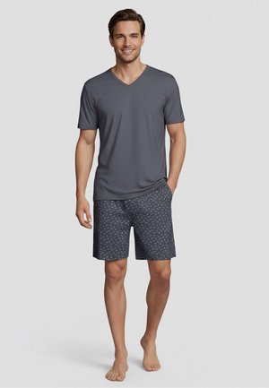 Glimlachende man die blootsvoets staat, draagt een grijs V-hals shirt met korte mouwen en een patroon van donkergrijze korte broek tegen een effen achtergrond.