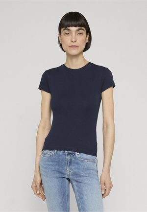 Femme aux cheveux courts et foncés portant un t-shirt bleu marine ajusté à manches courtes et un jean bleu clair, debout devant un fond uni.