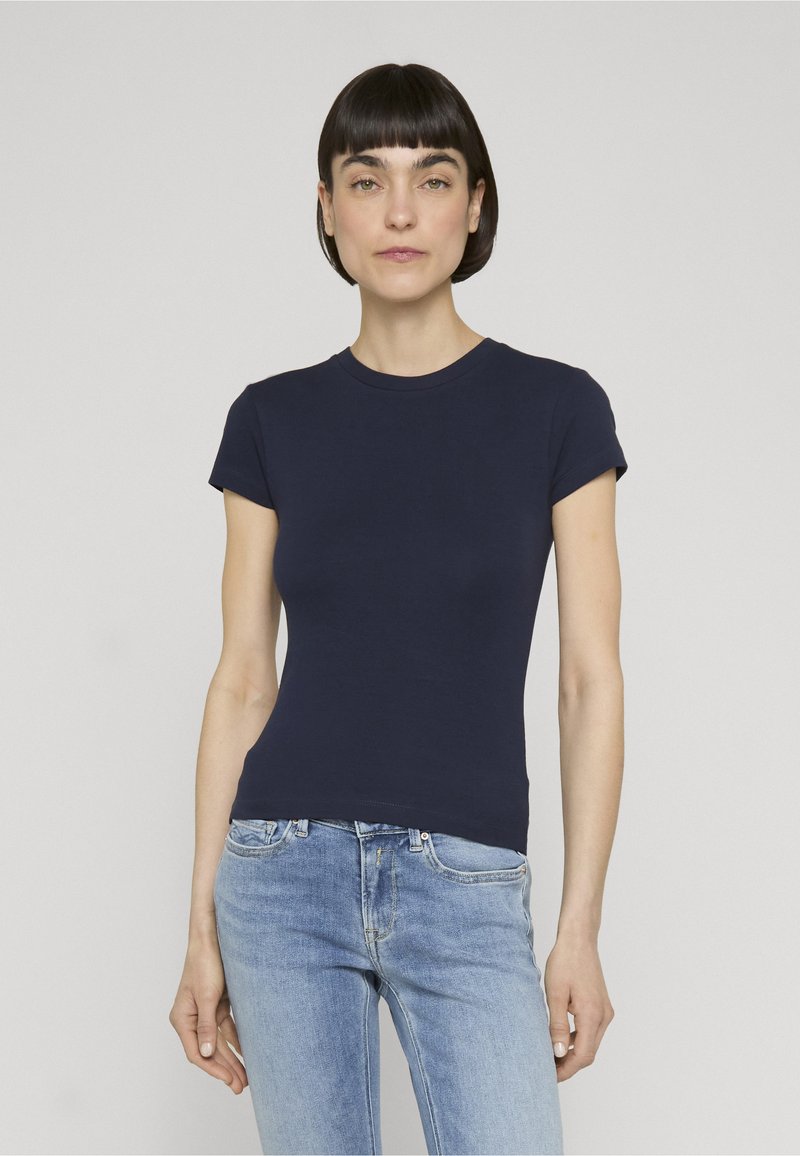 Femme aux cheveux courts et foncés portant un t-shirt bleu marine ajusté à manches courtes et un jean bleu clair, debout devant un fond uni.