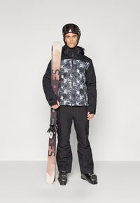 Echipament de schi care include o geacă neagră cu un model de camuflaj, pantaloni negri și o pereche de schiuri. Costumul include și o cască și ochelari de schi.
