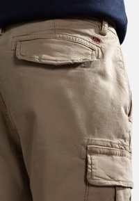 Beżowe spodnie cargo z boczną kieszenią, wykonane z miękkiej bawełnianej tkaniny o delikatnym połysku, z niewielkim haftem norweskiej flagi na tylnej kieszeni.