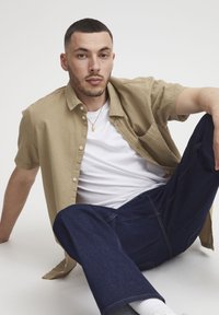 Korte mouwen, gestructureerd beige shirt met knopen, gecombineerd met een plain wit T-shirt en donkerblauwe jeans. Casual pose op een lichte achtergrond.