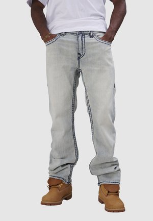 Pantalons en denim bleu clair avec des coutures foncées contrastantes, coupe décontractée et poches latérales, associés à des bottes de travail marron.
