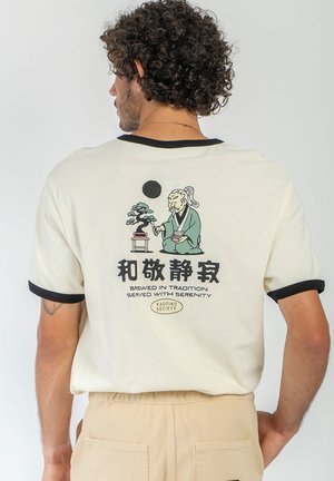 Mand med krøllet hår iført en cremefarvet t-shirt med sort kant, der har en japansk-inspireret illustration og tekst på ryggen.
