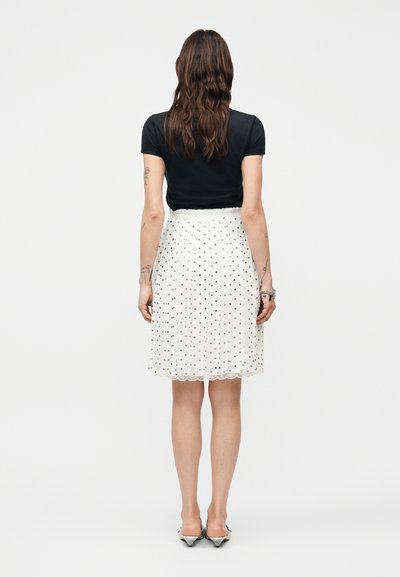 Γυναίκα με κυματιστά καστανά μαλλιά, φορώντας μαύρο τοπ με κοντά μανίκια, λευκή φούστα μέχρι το γόνατο με μαύρες βούλες και ασημένια μυτερά τακούνια, στέκεται με την πλάτη προς το μέρος μας.