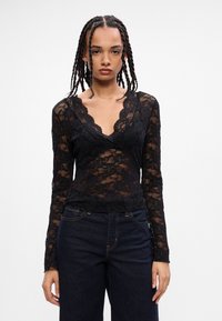 Zwarte kanten top met lange mouwen en een diepe V-hals, voorzien van geschulpte randen en bloempatronen, met een getailleerde pasvorm. Geweldig gecombineerd met donkere jeans.