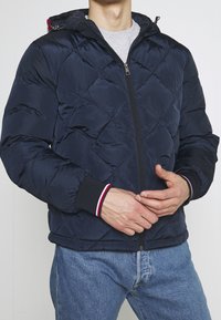 Man som bär en marinblå vadderad jacka med svarta ärmslut trimmade i röd, vit och blå, tillsammans med ljusblå jeans.