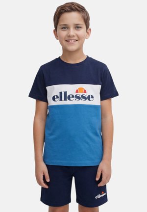 Ragazzo che indossa una t-shirt ellesse blu navy e blu e pantaloncini ellesse blu navy, in piedi e sorridente su uno sfondo semplice.