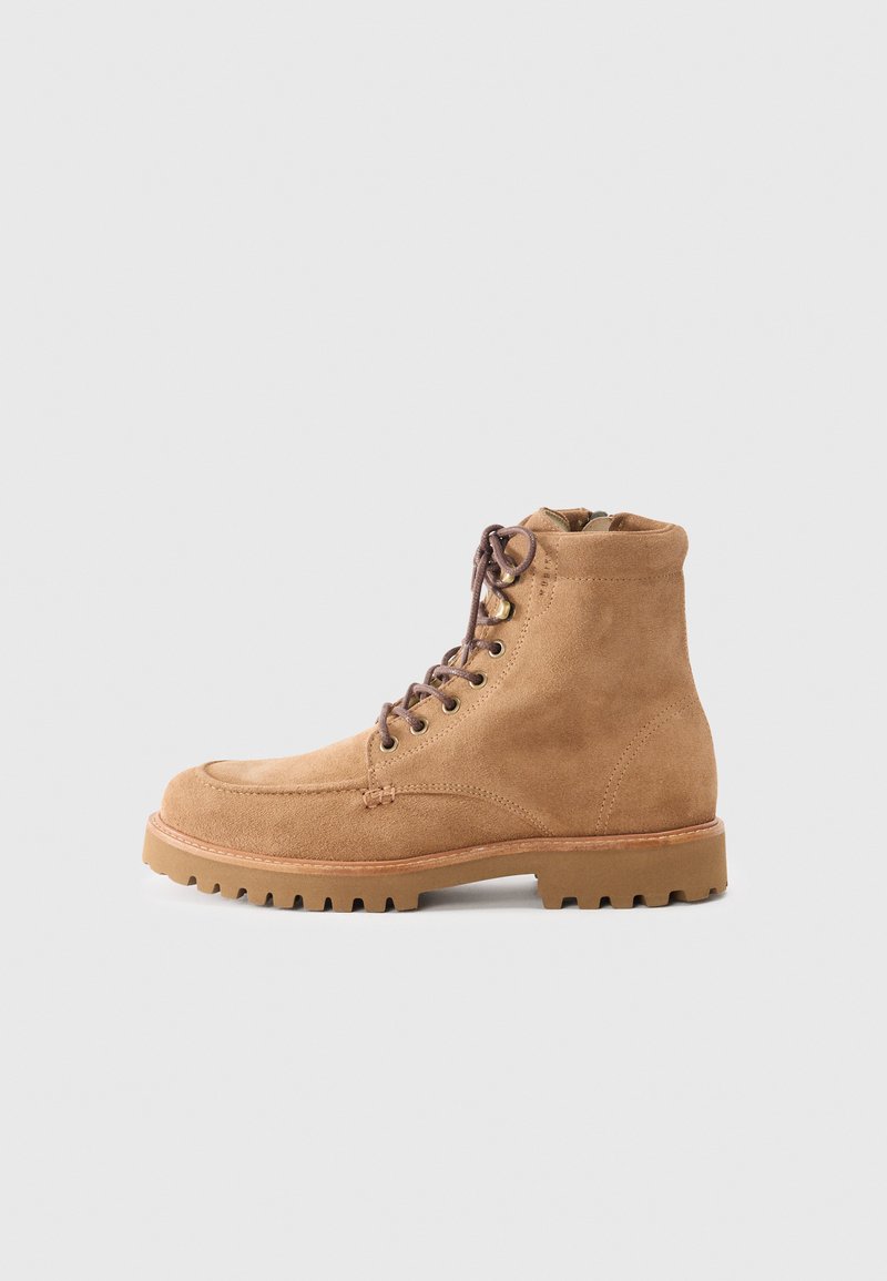Botine din suede bej cu vârful rotund, șireturi cu capse, tălpi rezistente și un finisaj texturat. Prezintă accente din hardware de culoare aurie.