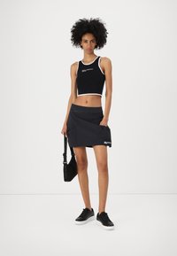 Fekete, rövidített tank top fehér díszítéssel, logóval, párosítva fekete szoknyával, oldalsó zsebekkel és húzózsinórral, fekete sportcipővel viselve.