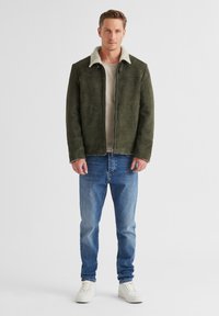 Olivgrüne Wildlederjacke mit einem weißen Kunstfellkragen, Reißverschluss und zwei Seitentaschen, kombiniert mit hellblauen Jeans und weißen Sneakers.