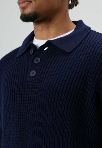 Close-up van een persoon die een marineblauwe gebreide trui draagt met een kraag en drie donkere knopen over een wit overhemd.