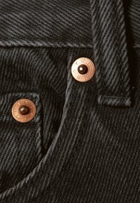 Tessuto di denim nero con trama sottile, caratterizzato da rivetti in rame con scritte in rilievo e una cucitura visibile.