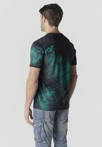 Mees, kes seisab seljaga, kannab musta ja rohelist plaastrimustriga T-särki ning helehalli sinist cargo-tüüpi põlvekaanetega teksapükse ühtlase tausta ees.