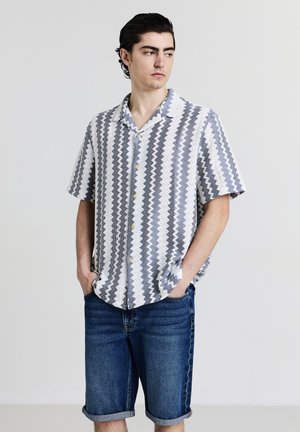 Young man with curly black hair wearing a blue and white zigzag short-sleeve shirt and rolled-up denim shorts, standing with hands in pockets.