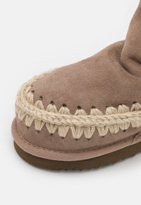 Botte en daim marron avec des accents tressés beiges, présentant un bout arrondi et une semelle texturée. Des coutures délicates soulignent le design.