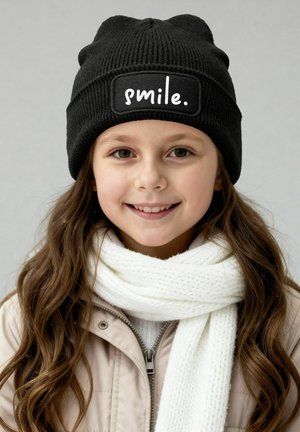 Schwarze Strickmütze mit einem weißen rechteckigen Aufnäher mit der Aufschrift "smile." Kombiniert mit einem weißen strukturierten Schal und einer beigen Jacke.