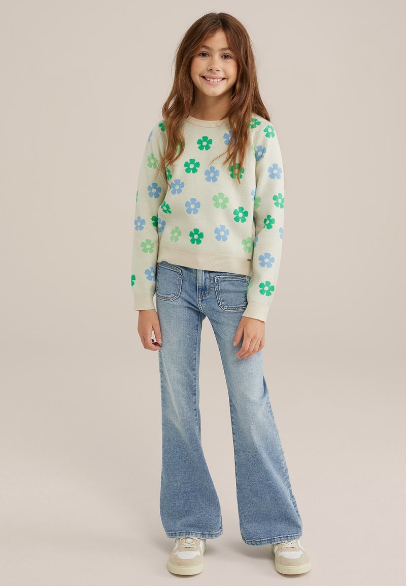 Ljust grönt tröja med blå och gröna blommönster, parat med utsvängda denimjeans. Modellen bär beige sneakers med vita detaljer.