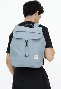 Ljusblå ryggsäck med lock, justerbara remmar och en liten logotypetikett. Tillverkad av hållbart tyg med en matt yta.