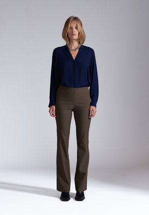 Frau steht frontal, trägt eine marineblaue Bluse, olivgrüne Hose, schwarze Schuhe und eine dicke silberne Halskette vor einem einfarbigen grauen Hintergrund.