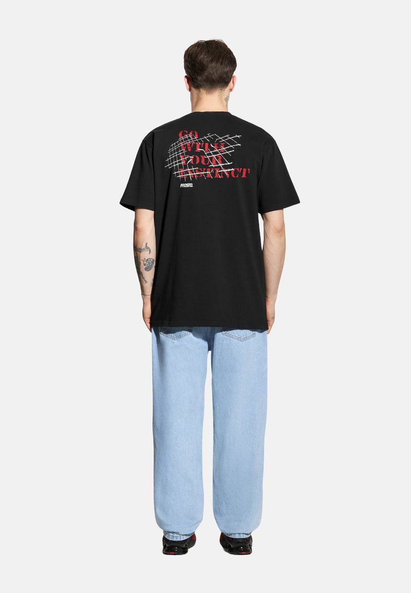 Czarna bawełniana koszulka z luźnym krojem. Na plecach znajduje się odważny czerwony i biały tekst oraz wzór w kratkę. Połączona z jasnoniebieskimi dżinsami.