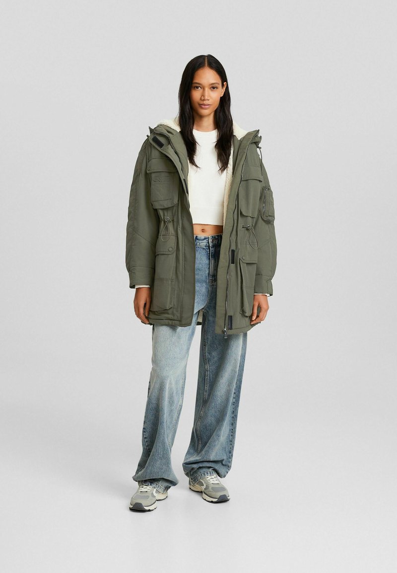Parka longue vert olive avec capuche en fourrure, plusieurs poches, taille cintrée, portée sur un crop top blanc et un jean bleu clair ample.