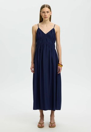 Femme aux cheveux lisses châtain clair portant une robe longue bleu marine sans manches et des sandales, debout sur un fond blanc.