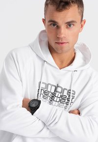 Witte hoodie met een grafische print in het zwart die "ombre menswear" leest. Een zwarte horloge met een ronde wijzerplaat wordt om de pols gedragen.