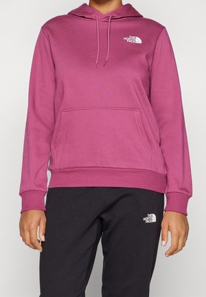 Osoba ubrana w różową bluzę z kapturem i czarne spodnie, oba z białymi logo The North Face na klatce piersiowej i udzie.