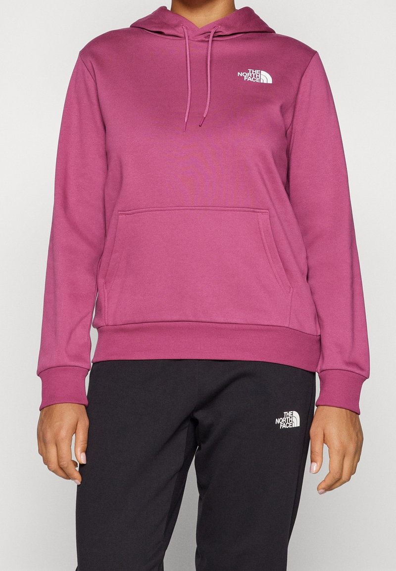 Personne portant un sweat à capuche rose et un pantalon noir, tous deux avec des logos blancs The North Face sur la poitrine et la cuisse.
