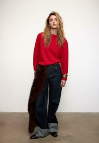 Maglione rosso, jeans di denim scuro larghi con un accento di pelliccia marrone su un lato e polsini di denim sfrangiati. Scarpe marroni e braccialetti d'oro.