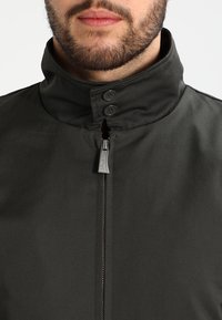 Ciemnoszara kurtka z kołnierzem, wyposażona w wysoki kołnierz, dwa guziki oraz przedni zamek błyskawiczny. Materiał ma gładką, trwałą fakturę.