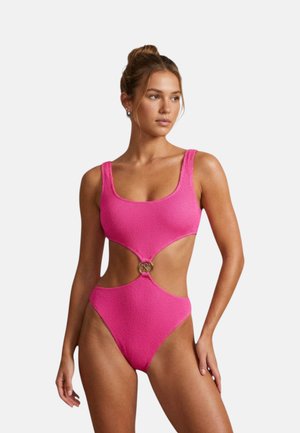 Donna con costume intero rosa acceso, strutturato con ritagli e dettaglio ad anello metallico sulla zona centrale, che guarda verso sinistra con i capelli raccolti in un chignon.