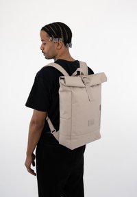 Roll-top backpack in light beige fabric with padded shoulder straps, side zipper pocket, and rectangular shape. Features a sturdy buckle closure.