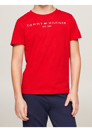 Enfant portant un t-shirt rouge Tommy Hilfiger avec logo et texte et un pantalon bleu marine, debout devant un fond uni.
