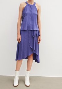 Lila ärmlös klänning med strukturerad vågig mönster, asymmetrisk nederkant och ett knytband i midjan, kombinerad med vita ankelskor.