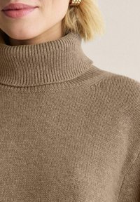 Ruskea, uritettu kauluspusero, joka on valmistettu pehmeästä neulomateriaalista. Lähikuva näyttää tekstuurin ja korkean kauluksen yksityiskohdat.