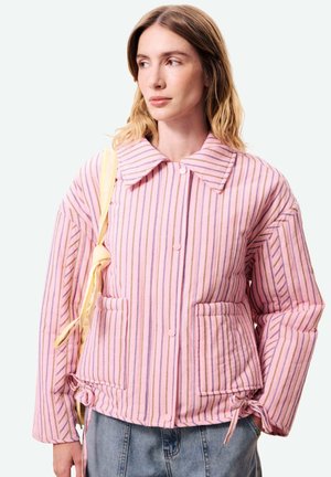 Vrouw draagt een roze gestreepte gewatteerde jas met grote kraag, voorzakken, gestrikte boorden en draagt een vaalgele tas over haar schouder.