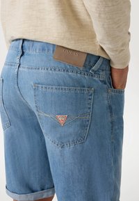 Svetlo modri denim kratki hlače z zavitimi robovi, zadnjimi žepi s šivi in rjavim usnjenim logotipom na pasu.