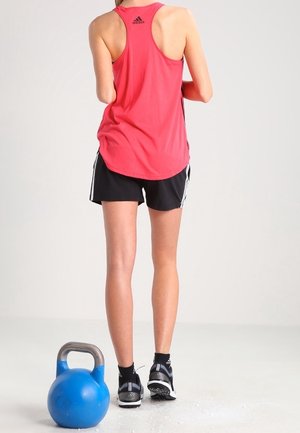 Top deportivo rojo sin mangas y pantalones cortos negros, acompañados de una kettlebell azul. Las zapatillas tienen detalles en negro y blanco. El fondo es liso y claro.